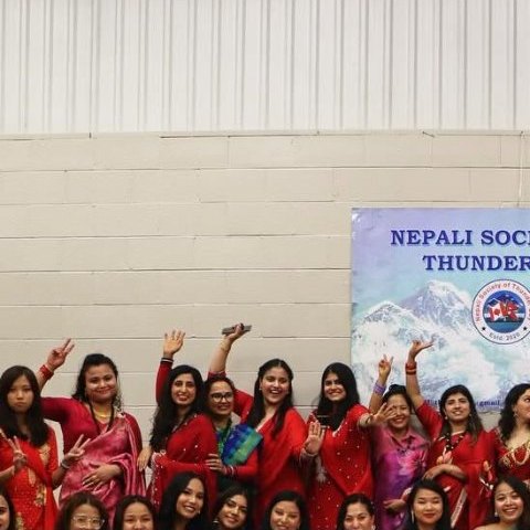A large group in red saris waving at the camera beneath the Nepali Society of Thunder Bay banner.